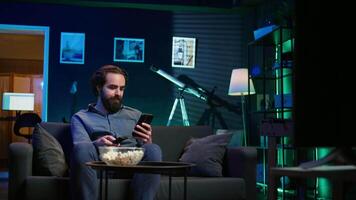 Smiling person on couch picking movie on streaming service using remote and texting on mobile phone. Happy man watching shows on television, zapping though channels, chatting smartphone, camera B video