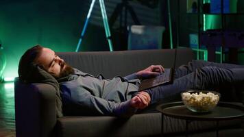 Exhausted man sleeping on couch after working on laptop all day before managing to watch TV and eat popcorn. Freelancer falls asleep while solving tasks on notebook and watching film, camera A video