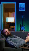 Vertical Amused man lounged on couch laughing at funny comedy movie while eating from bowl of popcorn. Cheerful person laying down in apartment amused by hilarious humor in film scene, camera B close up video