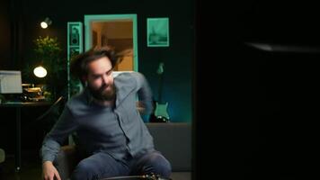 Cheerful gamer rushing home to play newly released game. Happy man jumping on couch, eager to play videogame on console, relaxing after hard day at work, camera B slow motion shot video