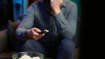 Close up of man in dimly lit apartment using remote control to browse though movies on streaming service. Remote control used by person in living room flipping through channels on TV, camera A video