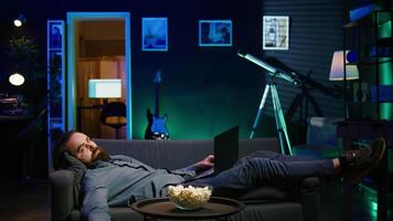 Exhausted man sleeping on couch after working on laptop all day before managing to watch TV and eat popcorn. Freelancer falls asleep while solving tasks on notebook and watching film, camera B video