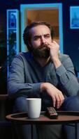 Vertical Man at home shocked by thriller film on TV, drinking coffee. Stupefied person at home left with eyes wide open by dramatic plot twist, watching suspenseful movie on TV, enjoying beverage, camera B video