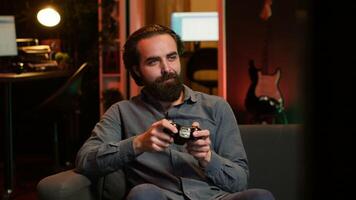 Joyful gamer at home gaming and enjoying leisure time. Smiling man sitting on couch in neon lit living room, excited to play videogame on console, relaxing after hard day at work, camera A video