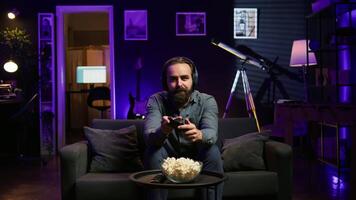Excited man playing videogames using controller, having fun in neon illuminated room. Enthusiastic gamer competing in multiplayer game using gamepad, talking with friends through headset, camera B video