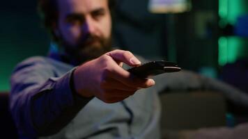 Close up of device used by person to scroll through streaming content, picking movie. Focus on remote control used by man in blurry background switching channels on television screen, camera A video