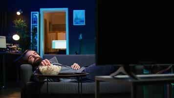 leende man lounged på soffa på Hem äter popcorn medan bläddring fastän bio tillgängliga på strömning service, njuter tid av från arbete. man om ner i lägenhet förbrukande mellanmål, kamera b video