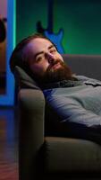 Vertical Smiling man lounged on couch eating popcorn while watching movie on streaming service, enjoying time off. Upbeat person laying down in apartment consuming snacks, looking at films, camera A video
