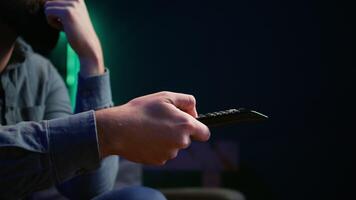 stänga upp av man på soffa använder sig av avlägsen kontrollera till bläddra fastän bio tillgängliga på strömning service. fokus på avlägsen kontrollera Begagnade förbi husägare i lägenhet växlande kanaler på tv, kamera b video