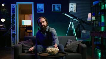 Man turning on TV using remote control, enjoying leisure time at home, munching on popcorn. Couch potato person in neon illuminated apartment eating salty snack, entertained by television, camera B video