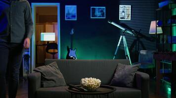 Man taking seat on sofa to watch television, getting entertained in relaxed home environment. Person arriving in apartment sitting down on couch to enjoy movies, turning on TV, eats popcorn, camera B video