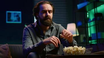 Lazy couch potato person mindlessly watching shows on TV and eating popcorn, paying close attention to screen. Sedentary man devouring snacks while consuming mindless entertainment, camera A video