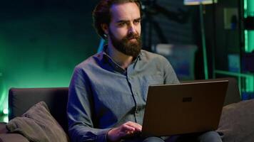 Man watching shocking breaking news story on TV while using laptop in neon lit apartment. Person turning volume on TV, surprised by events while sitting on couch using notebook, camera A video