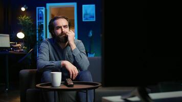 Man at home shocked by thriller film on TV, drinking coffee. Stupefied person at home left with eyes wide open by dramatic plot twist, watching suspenseful movie on TV, enjoying beverage, camera B video