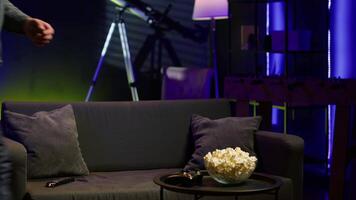 Gamer on couch in apartment engaging in online competitive multiplayer tourney using gamepad. Man in home theatre playing engaging videogame with controller, eating popcorn, camera A video