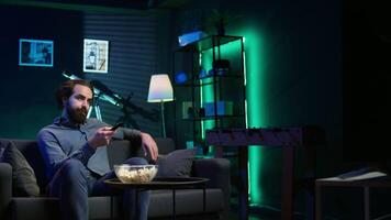 Man sitting on couch browsing though movies available on streaming service, enjoying time off from work. Person in apartment using remote control to switch channel on television set, camera B video