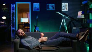 Smiling man lounged on couch eating popcorn while watching movie on streaming service, enjoying time off. Upbeat person laying down in apartment consuming snacks, looking at films, camera B video