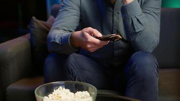 Close up of man on couch using remote control to browse though movies available on streaming service. Focus on remote control used by homeowner in apartment switching channels on TV, camera A video