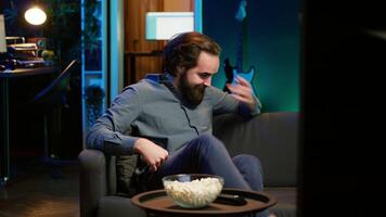 Amused man sitting on couch laughing while watching funny comedy movie, eating popcorn. Cheerful person in apartment consuming snacks and being amused by hilarious joke in film scene, camera A video