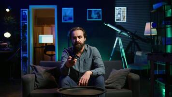 Bored person on couch in front of TV binging series on subscription based streaming services, browsing using remote. Weary man watching VOD shows on television set, zapping though channels, camera B video