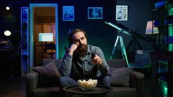 Bored man relaxing at home on couch, munching on popcorn while passing time watching movies on TV. Lazy person eating snacks and zapping through TV channels in RGB lit living room, camera B video