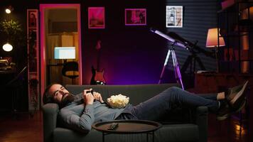 Gamer lounged on couch engaging in multiplayer game using gamepad, eating snack. Person in home theatre playing videogames on gaming console with bowl of popcorn in lap, camera B video