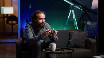 Focused man playing videogame in neon illuminated home, holding controller. Gamer hooked on enthralling game, excited to participate in online tournament, battling other players, camera A video