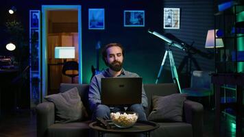 Man typing on laptop keyboard in neon lit apartment, sitting on couch and watching TV with bowl of popcorn ready. Teleworking person solving tasks on notebook with film playing in background, camera B video