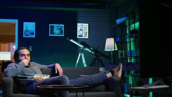 Homeowner bored at home munching on snacks, using TV to binge series on subscription based streaming services. Man sitting on couch, wasting time with VOD shows on TV, eating popcorn, camera B video