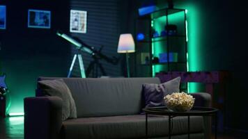 Bowl of popcorn on coffee table in front of cozy couch in empty neon lit modern apartment. Close up shot of snack prepared for movie night in home theatre used for entertainment sessions video
