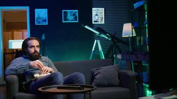 Man at home looking in disbelief at thriller film on TV, stuffing mouth with popcorn. Stupefied person at home watching suspenseful movie on VOD channel, perplexed by plot twist, camera B video