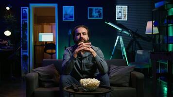 Man enjoying bowl of popcorn and smiling while watching cable programs on TV during leisure time at home. Spectator on couch in living room eating snacks and looking at television series, camera B video