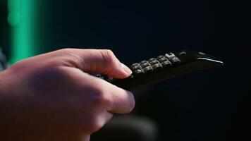 Close up of man in dimly lit apartment using remote control to browse though movies on streaming service. Remote control used by person in living room flipping through channels on TV, camera B video