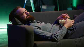 Amused man lounged on couch laughing at funny comedy movie while eating from bowl of popcorn. Cheerful person laying down in apartment amused by hilarious humor in film scene, camera A close up video