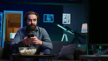 Man sitting on couch at home texting friend on smartphone, inviting him to watch movie on streaming service. Person at home discussing with mate on mobile phone, recommending TV show, camera B video