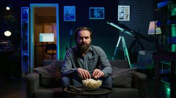 Man with surprised facial expression watching intriguing shows on TV and eating popcorn. Person paying close attention to screen, watching suspenseful thriller film, camera B close up video