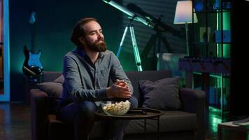 Man at home on couch gripped by interesting film shown on television screen. Person streaming content in neon lit living room, fascinated by entertaining movie scene, paying attention, camera A video
