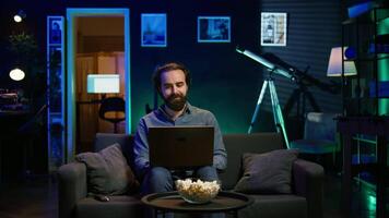 Man watching shocking breaking news story on TV while using laptop in neon lit apartment. Person turning volume on TV, surprised by events while sitting on couch using notebook, camera B video