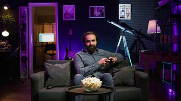 Cheerful gamer sitting on couch engaging in online competitive multiplayer tourney using gaming console and gamepad. Upbeat player engaging in fun videogames, enjoying leisure time, camera B video