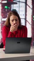 Vertical Tired woman sits at her desk staring at laptop screen and yawning in her loft, feeling overworked and exhausted. Person deals with burnout, stress and anxiety while juggling remote work. Camera B. video