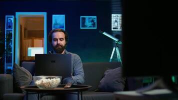 Man watching TV in apartment office and solving business related tasks on laptop, working from home. Freelancer on couch in living room looking at movies and working on notebook device, camera B video