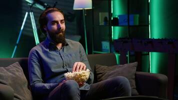 Amused person sitting on couch using TV to binge comedy series on subscription based streaming services, enjoying popcorn. Man laughing at funny shows on television, eating snack, camera A video