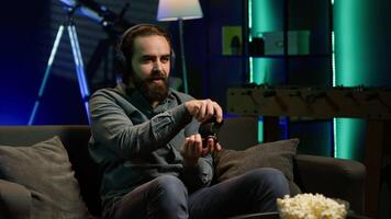 Gamer wearing headphones frustrated after getting game over message while playing videogame, raging. Man gaming with controller at home angry after losing level, camera A video