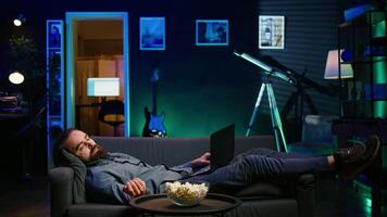 Sleepy man struggling to stay awake on couch after working on laptop all day before managing to watch TV and eat popcorn. Person falls asleep while solving tasks on notebook and watching film video