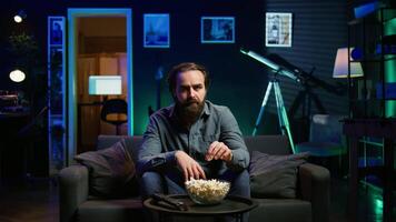 Puzzled man eating popcorn while watching confusing movie on streaming service, enjoying time off from work. Person consuming snacks, looking at film broadcasted on TV with convoluted plot, camera B video