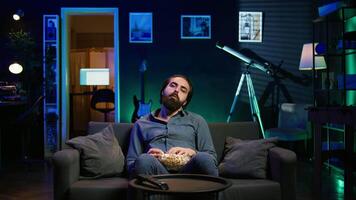 Fatigued man watching shows on TV late at night and munching on popcorn, paying close attention to screen. Tired sedentary man sitting in pajamas all day consuming mindless entertainment, camera B video