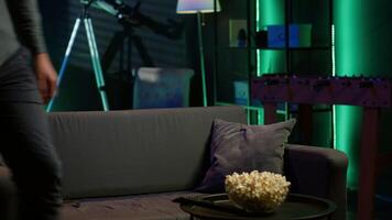 Man taking seat on sofa to watch television, getting entertained in relaxed home environment. Person arriving in apartment sitting down on couch to enjoy movies, turning on TV, eats popcorn, camera A video