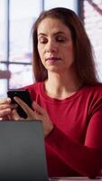 Vertical Focused woman in a home office loft talking on a mobile phone while working on laptop. She juggles calls, meetings and inquiries, using communication skills and creativity to solve tasks. Camera A. video