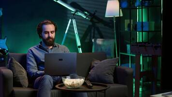 Man watching TV in apartment office and solving business related tasks on laptop, working from home. Freelancer on couch in living room looking at movies and working on notebook device, camera A video