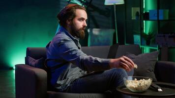 Man working on laptop lounged on neon lit apartment couch while picking TV channel to watch as distraction. Remote worker solving company tasks on notebook with TV show playing in background, camera A video